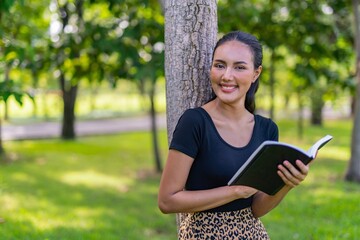 Portrait photo of the moment of a young beautiful asian female lady reading a book under a tree in an outdoor park during her summer holidays
