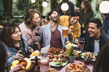 Multiracial group of hilarious friends have fun drinking beer and wine and eating together on the weekend at the restaurant table in the countryside