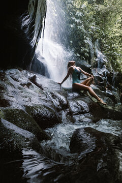 Woman in bikini sit on rock under falling water of Suwat waterfall in tropical jungle. Nature day tour, hiking activity adventure and fun at family tourist camp on summer vacation in Bali island