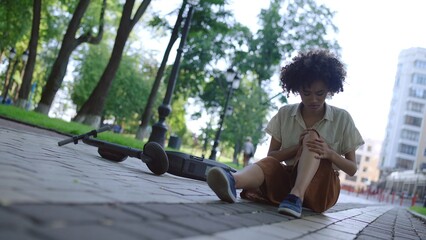 Young woman sitting on ground, rubbing injured knee after electric scooter accident