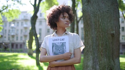 Upset African American woman looking around, holding missing cat poster in hands