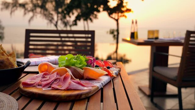 Romantic sunset dinner on the shore of a tropical island. Poolside Italian restaurant on beach. Table with luxurious food. Meat set with smoked pork, prosciutto. Served delicious antipasto in cafe.