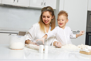 Obraz na płótnie Canvas Happy, funny, beautiful mom and son are messing around at a white table with dough in a white kitchen. Woman with a child have fun and fervently spend time together