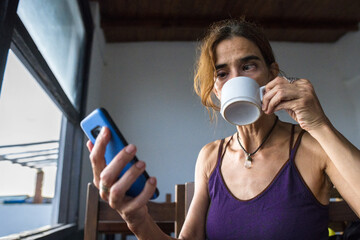 Latin adult female with squint eye working remote with mobile phone while drinking a latte espresso...
