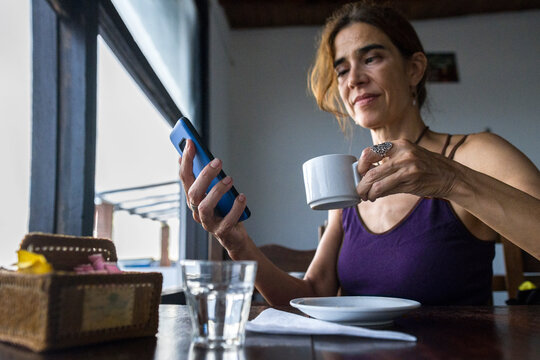 Latin Adult Woman With Strabismus Checking Her Mobile Phone And Drinking A Latte At Coffee Shop In The Morning. Remote Work Concept. Copy Space.