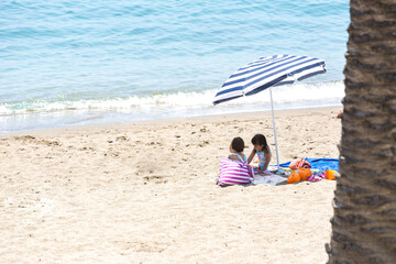 Twin sisters sitting on towels on the beach, under the shade of an umbrella, with a bag and armbands around them. Concept of beach, vacation, sun protection, sea, lifestyle and relaxation.