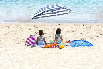 Twin sisters sitting on towels on the beach, under the shade of an umbrella, with a bag and...