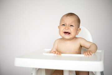 A small crying baby in a highchair. Whims. Refusal to eat. Crisis of 1 year. A light background, a place for text.