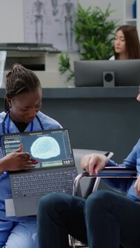 Vertical video: Nurse showing brain tomography to man with physical disability, doing medical consultation with patient wheelchair user. Looking at neurology scan diagnosis and neural system on laptop