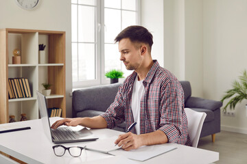 Concentrated young man working remotely on laptop and taking notes in notebook in home office. Serious man in casual clothes is sitting at table,typing on laptop and writing down necessary information