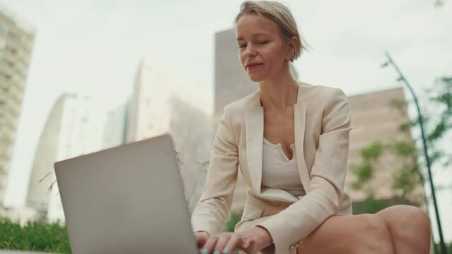 Smiling businesswoman with blond hair wearing beige suit is using pc laptop outside