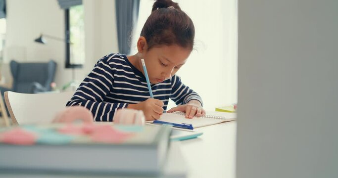 Asian toddler girl with sweater sit in front of desk with notepad use pencil focus on write notebook do homework from online learning course on the weekend at home. Distance online learning concept.