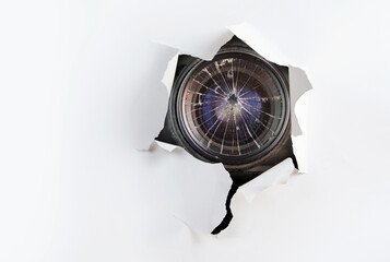 Concept of paparazzi or hidden camera, broken camera lens looks out through a hole in white paper wall