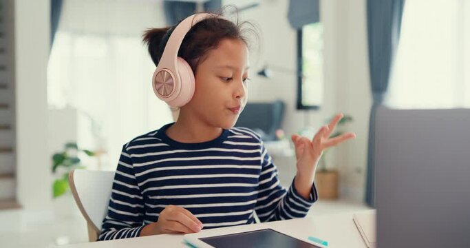 Asian toddler girl with sweater wear headphone sit front of desk with notepad use magic pen focus on writing do homework, finger counting number math online learning course from laptop at home.