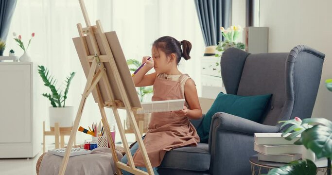 Asian toddler girl with apron sit on sofa chair in front of painting canvas with color palette use paint brush focus on painting abstract on weekend at home. Creative lifestyle for kid concept.