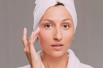 Young attractive woman in a bathrobe applies cream on her face in the bathroom. Close-up of the face.