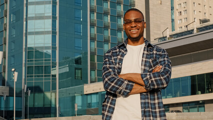 Stylish man in city african american guy young student male in sunglasses wears plaid shirt posing with crossed hands outdoors looking camera smiling toothy happy standing alone near company building