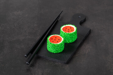 Obraz na płótnie Canvas Children Mousse cream cake with jelly watermelon in the form of round sweet Japanese sushi, sprinkled with green coconut chips, on a serving board. Light background. Copy space