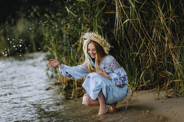 A beautiful, young happy blonde Ukrainian girl in an embroidered, long dress with a wreath of wheat and wild flowers sits near the sea and splashes water. Photography, Ukraine.