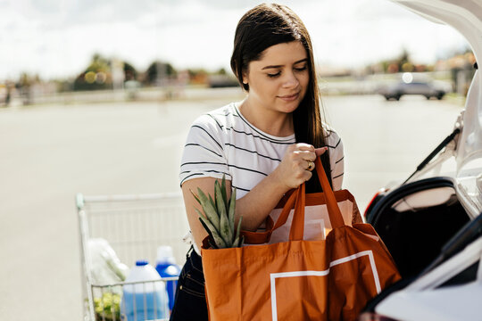 Young Woman Putting Package With Groceries In Car Trunk. Brazilian Woman Packing Groceries Into The Car Trunk