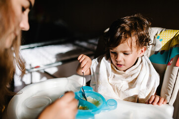 Mother feeding her baby girl with spoon. Mum giving healthy food to her adorable daughter at home. The girl eats porridge. Concept spoon-feeds.