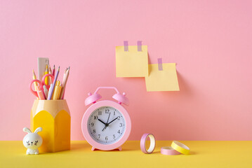 Back to school concept. Photo of school supplies pink alarm clock adhesive tape stand for pens bunny shaped sharpener and sticky note paper attached to pink wall - obrazy, fototapety, plakaty