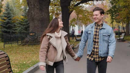 Cheerful two young spouses strolling on street in autumn park happy lovely couple enjoy pleasant walk holding hands talking carefree dreaming spend time together romantic date outdoors warm sunny day