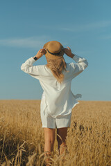Back view of young woman in a white dress and a straw hat on a wheat field in the summer.