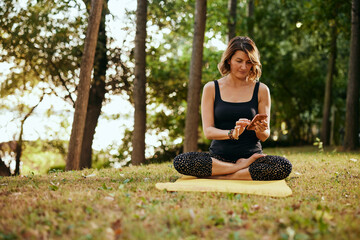 A yogi woman sits in a lotus pose and uses a smartphone in the forest.