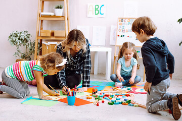 Young concentrated woman teacher sitting on floor with children, explaining how to draw with pencil in bright classroom.