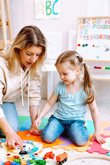 Little girl wear T-shirt, jeans sitting with young woman teacher teaching how to play with parts of clock with numbers.