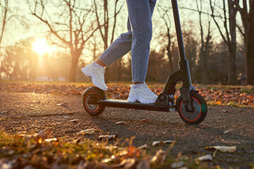 Woman discover city and park at sunset with electric scooter or e-scooter. Female Legs in sports sneakers stand on electric scooter. Girl riding on Ecological and urban transport.