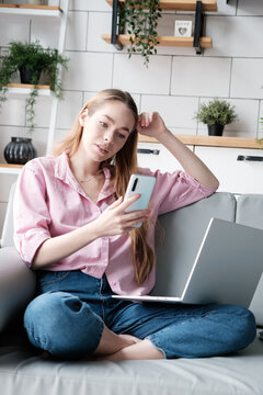 Attractive 20s aged caucasian young woman having video call with friend, smiling female student sits with laptop on sofa and chatting on mobile phone, blogger webcast live online, remote work at home
