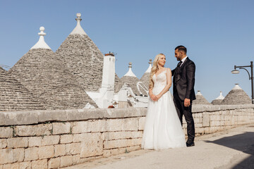 Happy wedding couple standing on street near conical roofs of trullo, meeting eyes. Young bride...