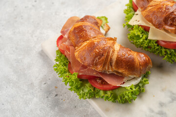 two lye croissant sandwiches with iberian ham, tomato slices, lettuce and cheese on marble board on grey background