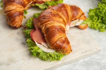 two lye croissant sandwiches with iberian ham, tomato slices, lettuce and cheese on marble board on grey background
