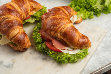 two lye croissant sandwiches with iberian ham, tomato slices, lettuce and cheese on marble board on...