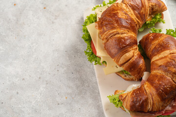 two lye croissant sandwiches with iberian ham, tomato slices, lettuce and cheese on marble board on grey background, space for text