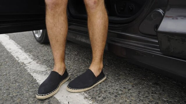 The Door Of A Parked Car Opens And A Caucasian Man Steps Out Of The Car. The Male Driver Gets Out Of The Car. A Guy In Light Summer Shoes Stands On An Asphalt Road With Road Markings