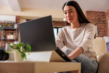 Asian authentic young female freelancer using laptop computer, communicates online web on internet with customer in cozy flat home apartment. Cozy home office workplace, remote work,E learning concept