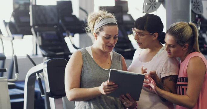 Talking female fitness group on a tablet browsing the web and social media at a gym. Women exercise group watching a video and talking together. Fitness class looking online after a training workout