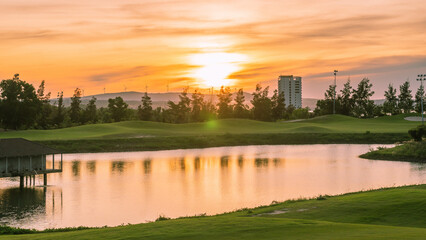 Orange red dawn sun above horizon, close-up lake pond foreground water texture surface calm reflection, trees high building background park city, hills green grass mountains, fabulously mysterious
