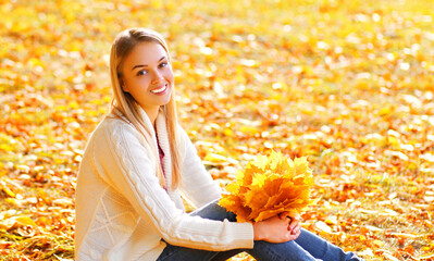 Portrait of beautiful blonde woman sitting with yellow maple leaves in autumn park