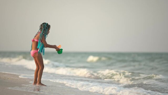 A girl with african braids in a summer costume plays on the beach with shells near the sea with waves under sunny sunset