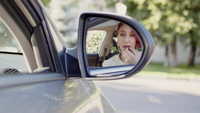 beautiful young redhead woman paints her lips with red lipstick in car while sitting in passenger seat and looking into side mirror. Applying make-up on way to work or business meeting