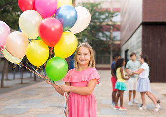 Fototapeta na wymiar Little blonde girl standing outdoor with multicolored balloons in hands. Her friends standing in background.