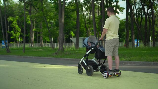 A young dad walks with a stroller on a hoverboard in the park. A modern dad on a gyro scooter walks with a child.