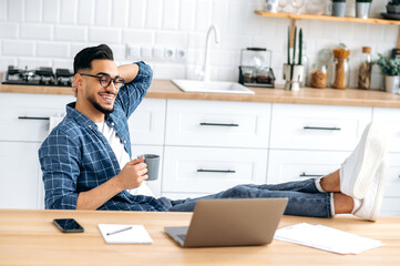 Relaxed indian or arabian modern guy in stylish clothes, sit at home in a kitchen with his legs on...