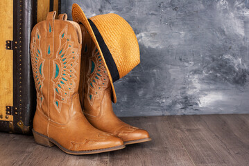 Leather cowgirl boots with straw hat pattern and an old suitcase stand on the floor against  gray...