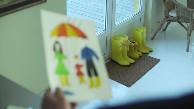 Father is looking at kids drawing of his family at home, children boots visible near entrance door.